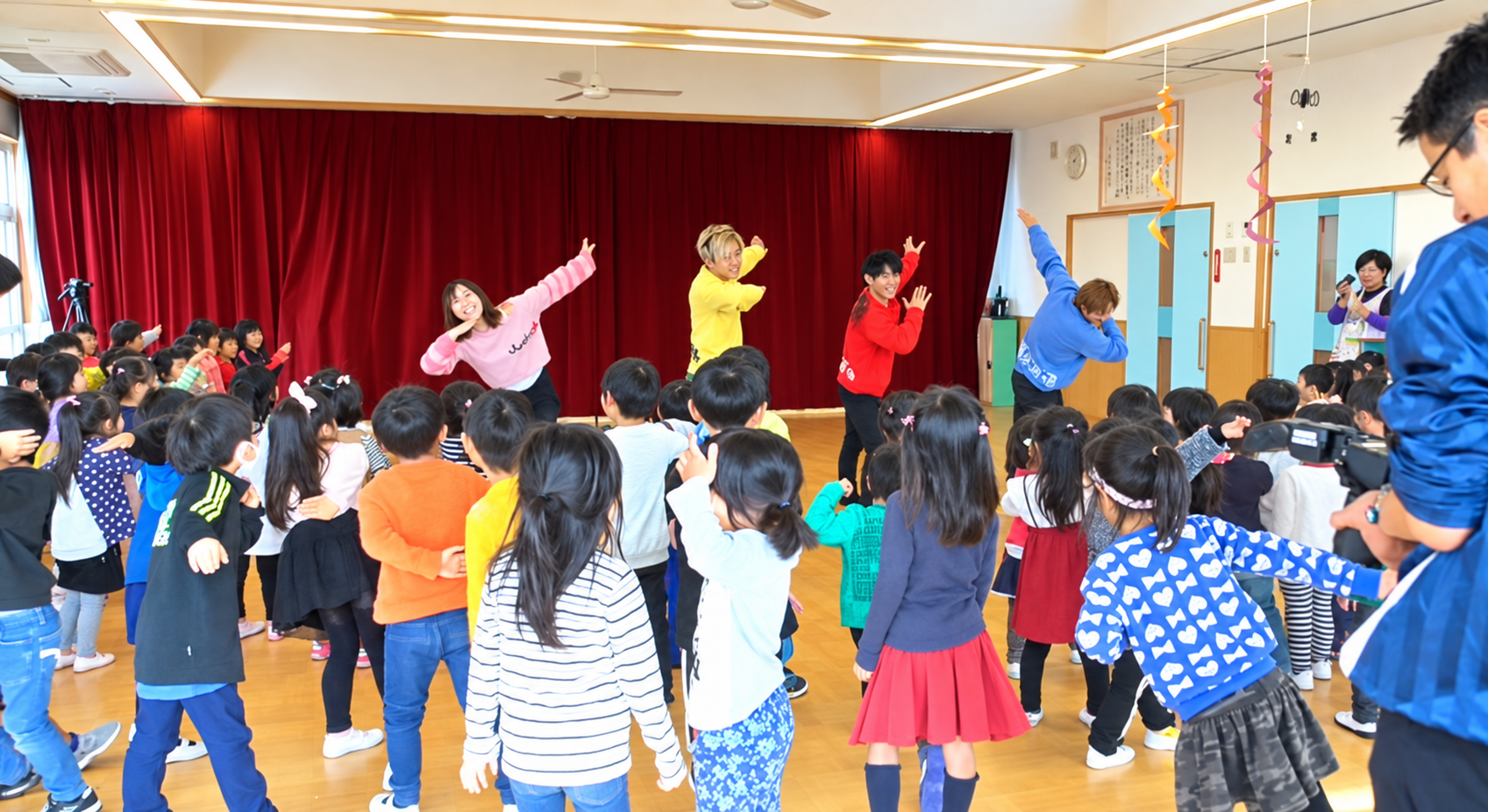 赤いカーテンの前で踊る4人の出演者と、その動きをまねる園児たち